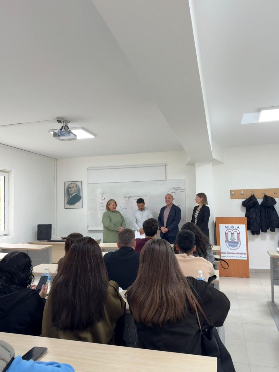 Bu fotoğraf, Muğla Sıtkı Koçman Üniversitesi'nde bir ödül töreni gibi bir etkinlik sırasında çekilmiş. Törende, ortada bir kişi ödül alırken, yanında üç kişi var. Sol taraftaki kişi, ödül alan kişiye gülümsüyor ve ödülü tutuyor. Sağ taraftaki diğer kişiler de ödül törenine katılan akademik yetkililer gibi görünüyor. Arka planda, duvarda dersin notları yazılı bir tahta bulunuyor.